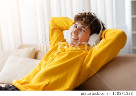Relaxed teen boy wearing wireless headphones and hoodie leaning back on sofa Relaxed teen boy wearing wireless headphones and hoodie leaning back on sofa 138571588