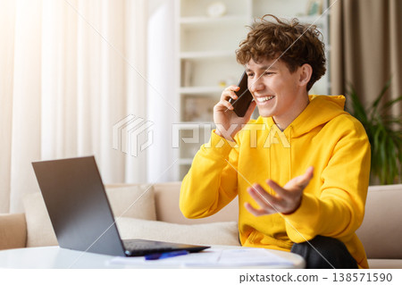 Happy young guy talking on phone with laptop and documents on table Happy young guy talking on phone with laptop and documents on table 138571590