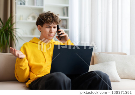 Confused frustrated teen boy in yellow hoodie on phone call with laptop Confused frustrated teen boy in yellow hoodie on phone call with laptop 138571591