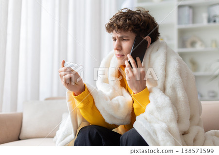 Sick teen boy wrapped in blanket talking on phone while holding thermometer Sick teen boy wrapped in blanket talking on phone while holding thermometer 138571599