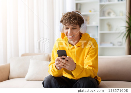 Young smiling teen guy with headphones sitting on sofa, looking at smartphone 138571608
