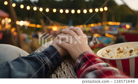 Close up of a romantic couple holding hands while enjoying popcorn at an outdoor movie festival with beautiful bokeh string lights during their relaxing summer vacation. AI Generated 138571649