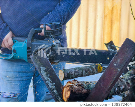 Man actively using an electric chainsaw to cut tree branches and logs into smaller pieces for firewood, working outdoors with sawdust flying and preparing wood for a home heating project. Man actively using an electric chainsaw to cut tree branches and logs into smaller pieces for firewood, working outdoors with sawdust flying and preparing wood for a home heating project. 138572404