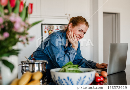 Mature woman searching recipe online on laptop while cooking in kitchen. Mature woman searching recipe online on laptop while cooking in kitchen. 138572619