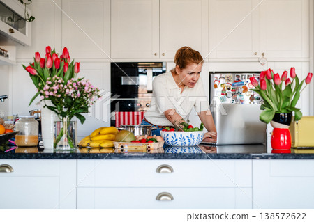 Mature woman searching recipe online on laptop while cooking in kitchen. Mature woman searching recipe online on laptop while cooking in kitchen. 138572622