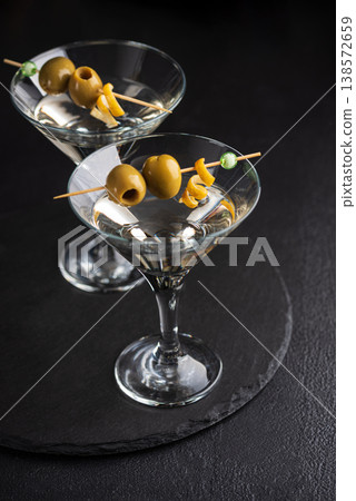 Two glasses with elegant cocktail martini with skewered green olives and a lemon twist on dark textured background. Two glasses with elegant cocktail martini with skewered green olives and a lemon twist on dark textured background. 138572659