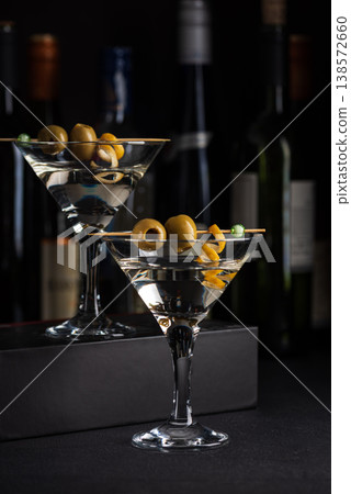 Two martini glasses with skewered green olives and citrus twists against dark surface with wine bottles blurred in the background. Two martini glasses with skewered green olives and citrus twists against dark surface with wine bottles blurred in the background. 138572660