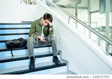 Depressed male student sitting on stairs and looking at phone. Depressed male student sitting on stairs and looking at phone. 138572783
