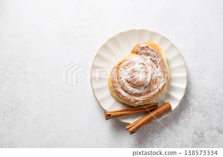 Freshly baked cinnamon roll with white icing and spice on a plate on a light background. Freshly baked cinnamon roll with white icing and spice on a plate on a light background. 138573334