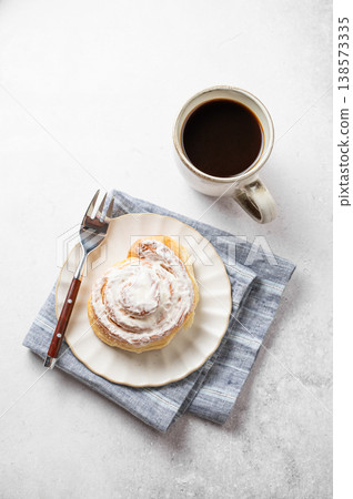 Freshly baked cinnamon roll with white icing and a cup of black coffee on a light background. Freshly baked cinnamon roll with white icing and a cup of black coffee on a light background. 138573335