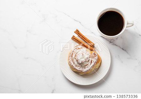 Freshly baked cinnamon roll with white icing and a cup of black coffee on a light marble background. Freshly baked cinnamon roll with white icing and a cup of black coffee on a light marble background. 138573336