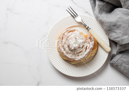 Freshly baked cinnamon roll with white icing on a plate, fork and napkin on a marble background. Freshly baked cinnamon roll with white icing on a plate, fork and napkin on a marble background. 138573337