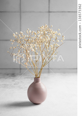 A dried bouquet of gypsophila flowers in a soft pink vase on a concrete table on a tile background. A dried bouquet of gypsophila flowers in a soft pink vase on a concrete table on a tile background. 138573362
