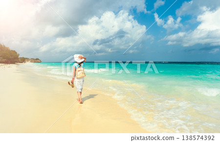 Beautiful white woman in long clothes walking along the beach, holding her hat 138574392