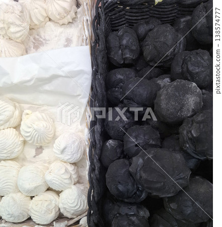 In a basket, there are white marshmallows and black marshmallows next to each other. Many people gather around to look at the unique shapes and colors during a busy market. In a basket, there are white marshmallows and black marshmallows next to each other. Many people gather around to look at the unique shapes and colors during a busy market. 138574777