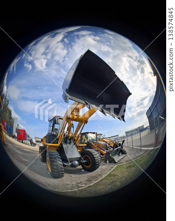 A yellow front loader stands with its bucket raised at a construction site. The bright cloudy sky creates a dramatic backdrop for the powerful machinery. A yellow front loader stands with its bucket raised at a construction site. The bright cloudy sky creates a dramatic backdrop for the powerful machinery. 138574845