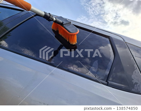 A person uses a long handled orange brush to clean the window of a silver car. The bright blue sky and white clouds reflect off the glass as they work in daylight. A person uses a long handled orange brush to clean the window of a silver car. The bright blue sky and white clouds reflect off the glass as they work in daylight. 138574850