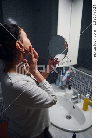Middle-Aged Woman in Pyjamas Checking Her Face in Bathroom Mirror During Morning Beauty Routine Middle-Aged Woman in Pyjamas Checking Her Face in Bathroom Mirror During Morning Beauty Routine 138575024