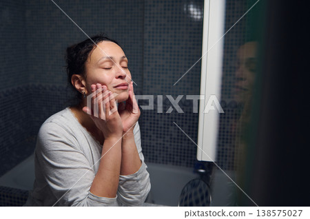 Middle-Aged Woman in Pajamas Doing Morning Skincare Routine at Bathroom Mirror 138575027