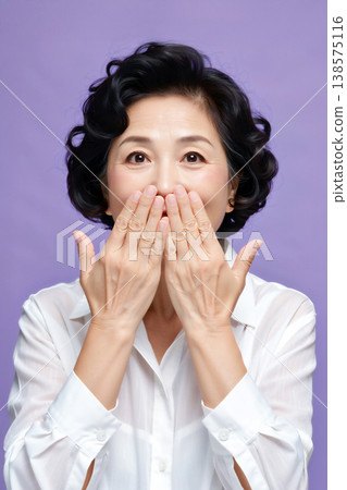 A senior woman smiles and covers her mouth with both hands. 138575116