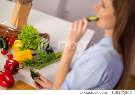 Waist up of a young woman eating a slice of cucumber in the kitchen Waist up of a young woman eating a slice of cucumber in the kitchen 138575207