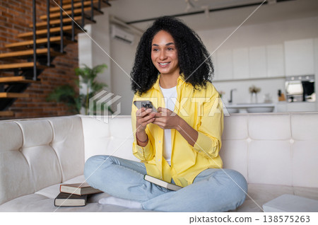Curly-haired young woman sitting on the sofa and texting on the phone Curly-haired young woman sitting on the sofa and texting on the phone 138575263