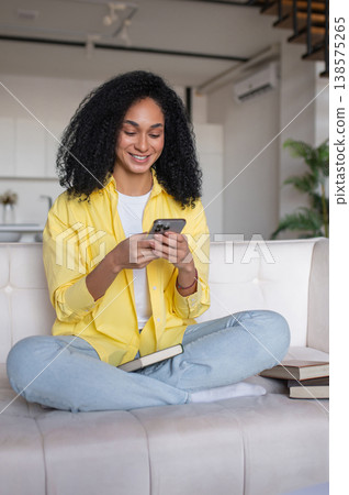 Curly-haired young woman sitting on the sofa and texting on the phone 138575265