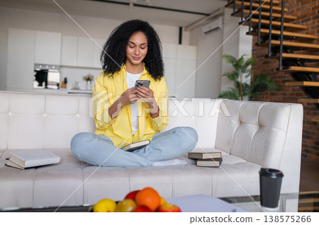 Curly-haired young woman sitting on the sofa and texting on the phone Curly-haired young woman sitting on the sofa and texting on the phone 138575266