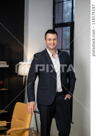 Male executive smiling and standing with hands in pockets in office setting Male executive smiling and standing with hands in pockets in office setting 138576197
