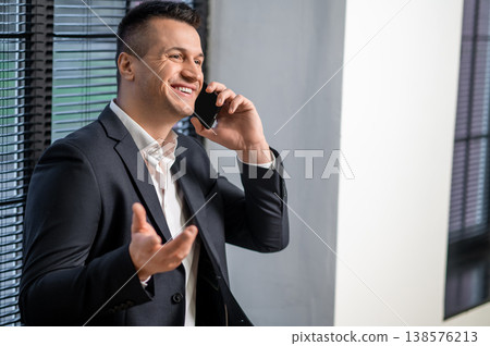 Businessman in suit laughing during phone call while posing in office Businessman in suit laughing during phone call while posing in office 138576213