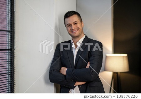 Smiling businessman standing near window with crossed hands 138576226