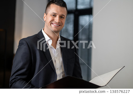 Businessman in suit smiling at camera holding report folder in office Businessman in suit smiling at camera holding report folder in office 138576227