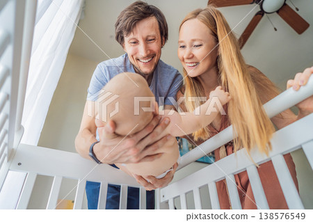 Mother and father lifting baby from crib and holding in arms at home. Loving parents caring for newborn together. Family bonding, parenthood, tenderness and emotional connection concept 138576549