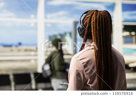 African american girl listening to music and waiting for boarding plane, walking around the airport terminal lounge area. Teenager preparing for international flight departure, diverse travelers. African american girl listening to music and waiting for boarding plane, walking around the airport terminal lounge area. Teenager preparing for international flight departure, diverse travelers. 138576918