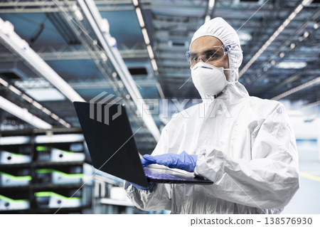 Man working in PCB production factory oversees infrastructure with laptop. Microchips manufacturing plant engineer in cleanroom tracking semiconductor fabrication lots with notebook 138576930