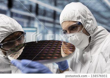 Fabrication engineers checking silicon wafer hardware defects in industry 4.0 manufacturing plant. Specialists in bunny suits assessing smart factory semiconductor substrate yields, close up 138576931