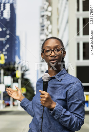 Field reporter holds mic, speaking towards camera on city street, presenting information. African american journalist reports live news update with microphone from metropolitan downtown area avenue 138576944
