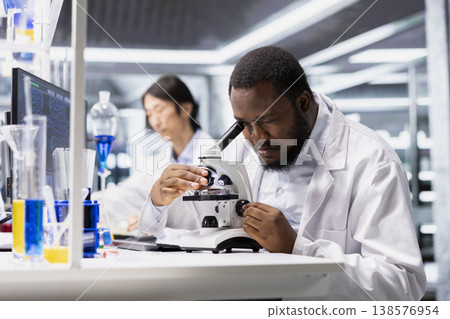 Materials scientist using microscope to evaluate cellular structures in lab workspace. African american man observes specimens through magnification tool at laboratory bench workstation 138576954