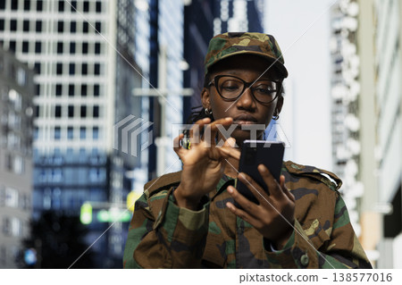 Army soldier examines smartphone map app while walking on city street. Female military officer using phone navigation application to check tactical route coordinates in urban environment Army soldier examines smartphone map app while walking on city street. Female military officer using phone navigation application to check tactical route coordinates in urban environment 138577016
