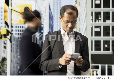 Indian male professional checking his mobile phone apps on crowded street, walking along busy avenue with a big crowd of diverse pedestrians. Adult on his morning commute downtown. Indian male professional checking his mobile phone apps on crowded street, walking along busy avenue with a big crowd of diverse pedestrians. Adult on his morning commute downtown. 138577019