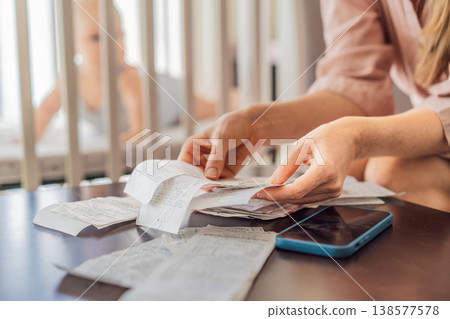 Mother sitting on sofa calculating monthly expenses with receipts while baby lies in crib nearby at home. Stressed mom managing budget and childcare. Financial pressure and parenthood concept Mother sitting on sofa calculating monthly expenses with receipts while baby lies in crib nearby at home. Stressed mom managing budget and childcare. Financial pressure and parenthood concept 138577578