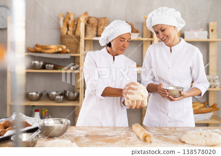 Two skilled female bakers in white uniform kneading and cutting dough, preparing portioned pieces before baking in bakehouse Two skilled female bakers in white uniform kneading and cutting dough, preparing portioned pieces before baking in bakehouse 138578020