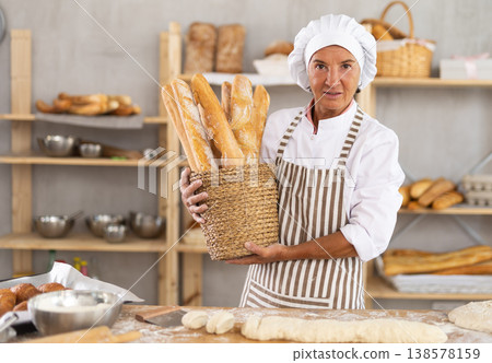 Happy mature female baker posing with basket of freshly baked baguettes in bakery, demonstrating concept of food industry and baker profession 138578159