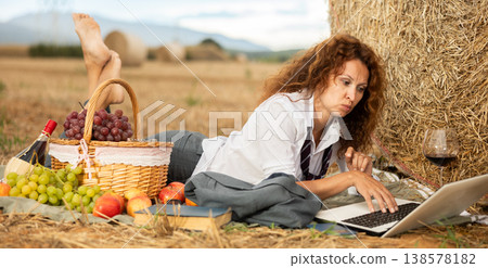 Woman in white blouse in office clothes, lying on blanket and working on laptop. Picnic on farm field Woman in white blouse in office clothes, lying on blanket and working on laptop. Picnic on farm field 138578182