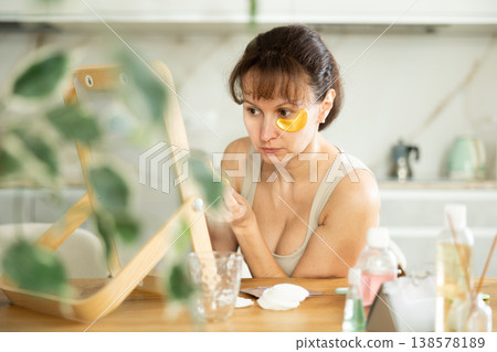 Middle-aged woman applying eye patch mask on her face sitting in front of the mirror Middle-aged woman applying eye patch mask on her face sitting in front of the mirror 138578189