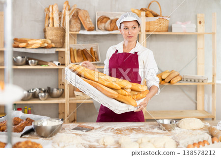 Successful female baker holding wicker tray of fresh baguettes Successful female baker holding wicker tray of fresh baguettes 138578212