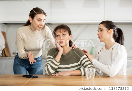 Sad woman sits silently in kitchen, adult girls sympathize and comfort her 138578213