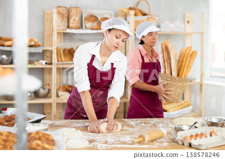 Two professional female baker in apron working in bakehouse. Young woman kneading dough and mature woman arranging freshly baked bakery products 138578246