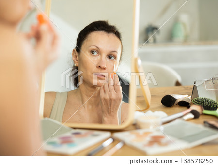 Middle-aged woman applying concealer on her facial skin sitting in front of the mirror Middle-aged woman applying concealer on her facial skin sitting in front of the mirror 138578250