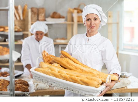 Successful female baker holding wicker tray of fresh baguettes Successful female baker holding wicker tray of fresh baguettes 138578253